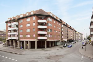Flerbostadshus i röd tegel vid en korsning, med balkonger, butikslokaler i bottenvåningen och trottoarer längs gatan