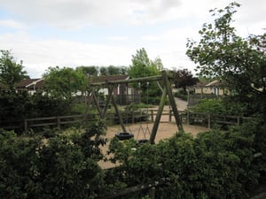 Liten lekplats med gungor omgiven av gröna buskar och träd, med hus i bakgrunden