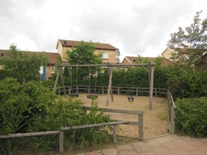 Lekplats med gungor omgiven av grönska och staket, med bostadshus i bakgrunden under en mulen himmel