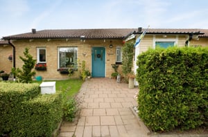 Ett enplanshus med gul tegelfasad, blå ytterdörr och en trädgård med buskar och blommor