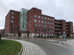 Bostadshus i tegel med flera våningar, omgivna av trottoarer, väg och grönområde under en molnig himmel