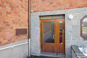 Entrédörr i trä med glaspanel i en tegelfasad, omgiven av stenramar och en belysningsarmatur på väggen