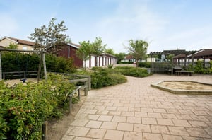 Lekplats med gungställning, sandlåda och omgivande grönska, belägen nära röda byggnader under en ljus himmel