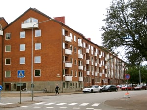 Flerbostadshus i rött tegel med vita balkonger, parkerade bilar och en trottoar i förgrunden. En person går över ett övergångsställe nära ett trafikskyltat område