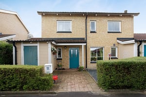 Tvåvåningshus med tegelväggar, blå ytterdörr och fönster med vita ramar, omgiven av gröna häckar och en stenlagd gång