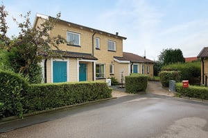 Tvåvåningshus med gul tegelfasad, blå dörrar och gröna häckar runt tomten, beläget vid en asfalterad väg under klar himmel