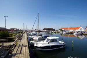 Båtar förtöjda vid en liten hamn med brygga, omgivna av byggnader och klarblå himmel