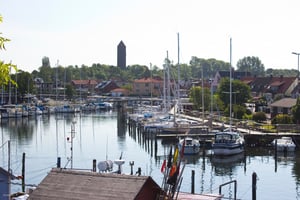 Småbåtshamn med segelbåtar och motorbåtar vid kaj, omgivna av hus och grönska med en kyrktornliknande byggnad i bakgrunden