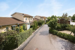 Rad av moderna radhus med tegeltak längs en asfalterad gata, omgiven av grönskande häckar och träd under en klar blå himmel