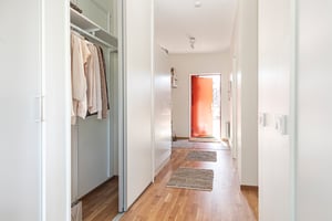 Hall med ljusa väggar, trägolv, öppna garderober och en röd ytterdörr med mattor på golvet