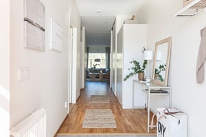 Ljus hall med trägolv, vita väggar, och inredning inklusive spegel, växt, och förvaringsmöbler. Vy mot vardagsrum med stora fönster i bakgrunden