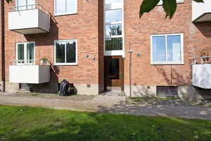 Tegelbyggnad med röda fasader, vita balkonger och glasdörr vid ingång. Gräsmatta och stenlagt gångområde framför byggnaden