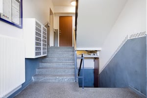 Trapphus med gråa golv, vita väggar och en trappa som leder både uppåt och nedåt. Dörrar, brevlådor och radiator syns på bilden
