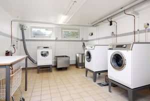 Tvättstuga med tre tvättmaskiner, torktumlare, och en arbetsbänk i ett ljust rum med klinkergolv och vita kaklade väggar