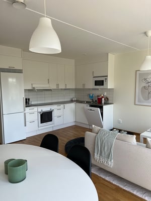 Modernt kök och vardagsrum i öppen planlösning med vita skåpluckor, matbord med svarta stolar och grå soffa med filt