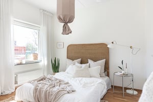 Ljust sovrum med en säng med rottinggavel, vita sängkläder, ett fönster med vita gardiner, en grön växt, och en golvlampa