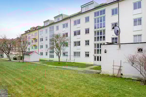 Flerbostadshus med vit fasad, grön gräsmatta och träd i förgrunden under en klar himmel