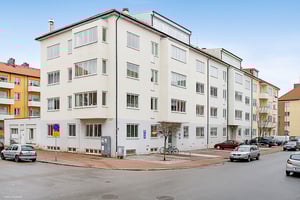 Vit flervåningsbyggnad med stora fönster, belägen vid en trottoar intill en mindre gata med parkerade bilar