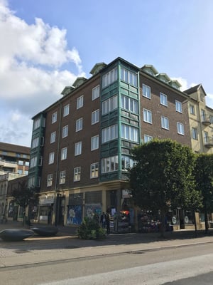 Flerbostadshus med tegelfasad och gröna detaljer, omgiven av träd och en trottoar under en blå himmel med moln