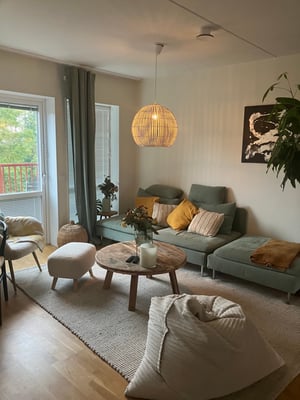 Vardagsrum med grå soffa, soffbord i trä, plädar och prydnadskuddar. En hängande lampa i rotting och utsikt mot en balkong med grönska