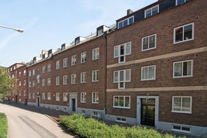 Byggnad med fasad i rödbrunt tegel, flera fönster samt vita detaljer, belägen vid en asfalterad gata och omgiven av grönska