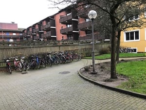 Cykelställ med parkerade cyklar på en stenlagd gångväg, omgiven av träd och flerbostadshus i bakgrunden