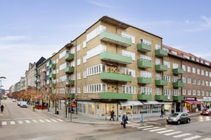 Flerbostadshus i tegel med gröna balkonger, butiker på bottenvåningen och trottoarer längs en gata med promenader och trafik