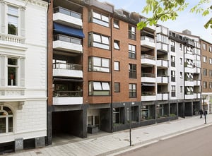 Flerbostadshus med fasad av tegel och puts, balkonger och stora fönster längs en trottoar i stadsmiljö