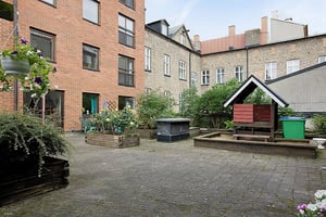 Innergård med stenlagd mark, växter i blomlådor, möbler och ett litet lekhus omgiven av byggnader