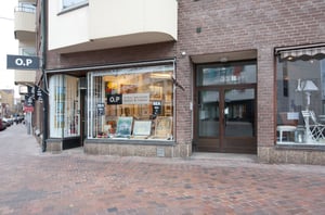 Fasad av en butik med skyltar som säger "O.P" och "REA", bredvid en dörr med adressnumret 33. Tegelvägg och stenbelagd trottoar runt omkring