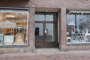 Butiksfasad med brun tegelvägg, två fönster och en glasdörr med nummer 33. Fönstret till vänster visar konstverk och en rea-skylt, medan fönstret till höger innehåller vita möbler