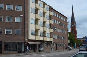 Flerbostadshus med beige balkonger och brun fasad vid en gata, med en kyrka med spetsigt torn i bakgrunden