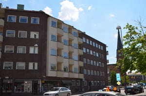 Flerbostadshus i brun och beige fasad med parkerade bilar på gatan, en kyrkobyggnad synlig i bakgrunden under en klarblå himmel