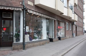 Fasad av en butik med stora glasfönster, tegelväggar och markis, belägen vid en trottoar och ett flerbostadshus