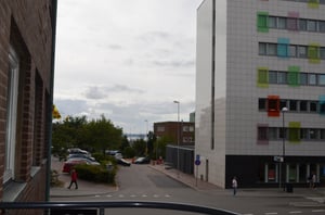 Vy över en stadsgata med parkerade bilar, träd och ett modernt byggnadskomplex med färgglada fönster. En person promenerar på trottoaren