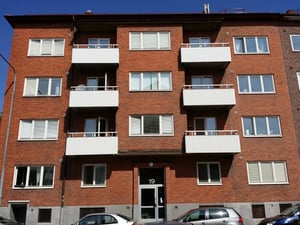 Flerfamiljshus med fasad av rött tegel, vita balkonger och ett flertal fönster. Parkerade bilar framför byggnaden
