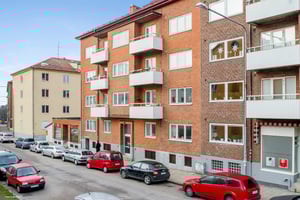 Gatubild med flera flerbostadshus i tegel, parkerade bilar längs trottoaren och klar blå himmel i bakgrunden