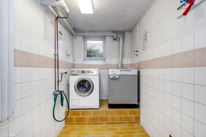 Tvättstuga med vit tvättmaskin och grå torktumlare placerade på gulbrunt klinkergolv. Väggarna är kaklade med vita och beige plattor