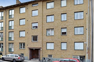 Gul tegelbyggnad med flera våningar, fönster och en entrédörr. Parkerade bilar och en cykel syns framför fasaden