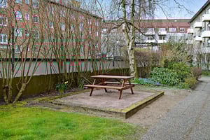 Grön parkmiljö med en röd träbord-och-bänksats på en stenlagd yta, omgiven av gräs, träd och buskar, med bostadshus i bakgrunden