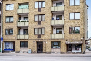 Gul tegelbyggnad med flera våningar, balkonger och butiken "Tapetserarverkstad" på bottenvåningen. En gata med parkeringsskylt syns i förgrunden