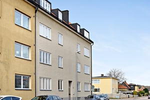 Flerfamiljshus med beige fasad, parkerade bilar längs gatan och ett gult hus i bakgrunden under en klar himmel