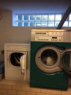 Tvättstuga med två tvättmaskiner i vitt och grönt, ståendes bredvid varandra i ett rum med kaklat golv och glasblocksvägg