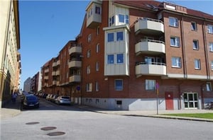 Flerfamiljshus i tegel med balkonger längs en gata, parkerade bilar och trottoarer på båda sidor