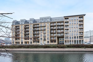 Flerfamiljshus vid vattnet med modern arkitektur och stora fönster, omgiven av träd och en blå himmel