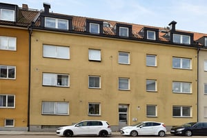 Gul flerfamiljsbyggnad med tre våningar och bruna takpannor, flera fönster och parkerade bilar framför