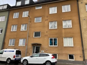 Gul flerfamiljsbyggnad med flera våningar, vit skåpbil och vit personbil parkerade framför