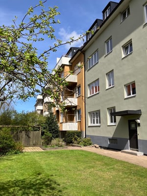 Flerfamiljshus med ljusa fasader och grönskande trädgård i förgrunden under en blå himmel