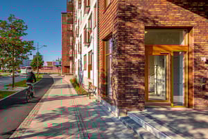 Flerfamiljshus i tegel med glasentré, cyklist på gång- och cykelväg, och träd i soligt stadslandskap