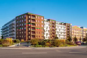 Flerfamiljshus med moderna fasader i tegel och ljusa färger, omgivna av grönska och en asfalterad väg i förgrunden